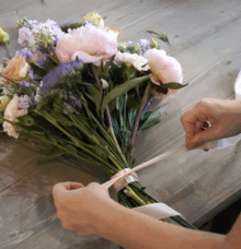En person binder ett band runt en bukett med blandade blommor på ett träbord.